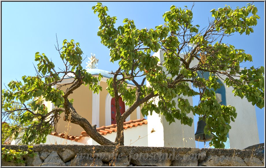 Zia_Tigaki_Fotoschule_010.jpg - Eine kleine weiß getünchte Kapelle mit kraftvoll blau abgesetzten Verzierungen, mitten auf einem gemütlichen Dorfplatz, auf dem ein großer Baum Schatten spendet, sorgt für eine unvergleichliche Stimmung.